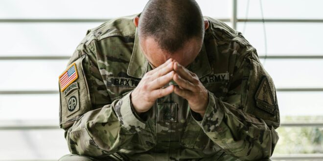 A soldier in camouflage sitting indoors, appearing stressed and overwhelmed.